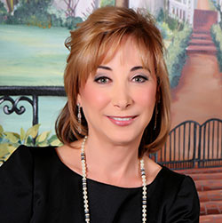 A woman with shoulder-length light brown hair, wearing a black top and a beaded necklace, poses in front of a painted outdoor background.