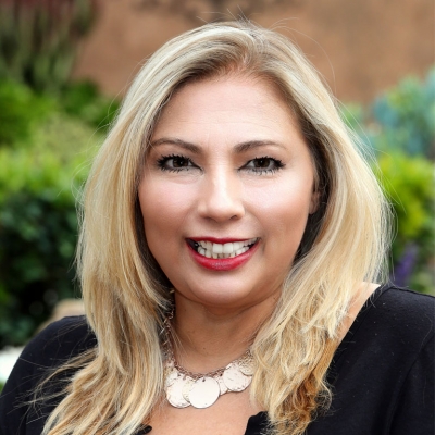 A woman with long blonde hair, wearing a black top and a metallic necklace, smiles while standing outdoors with greenery in the background.