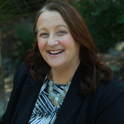 A woman with long brown hair wearing a black blazer and patterned top smiles outdoors, with greenery in the background.