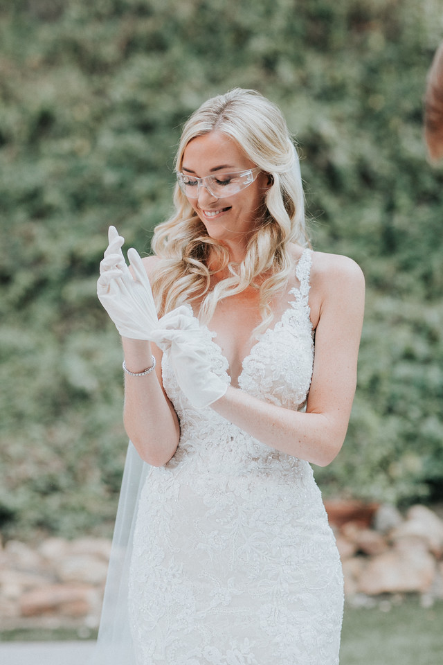 ceremony 154 X2 A bride in a white lace wedding dress and safety glasses puts on white gloves outdoors, with greenery in the background.