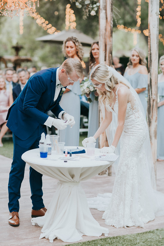 ceremony 172 X2 A bride and groom, both in formal attire and wearing gloves, mix chemicals at a table during an outdoor wedding ceremony, with bridesmaids and guests in the background.