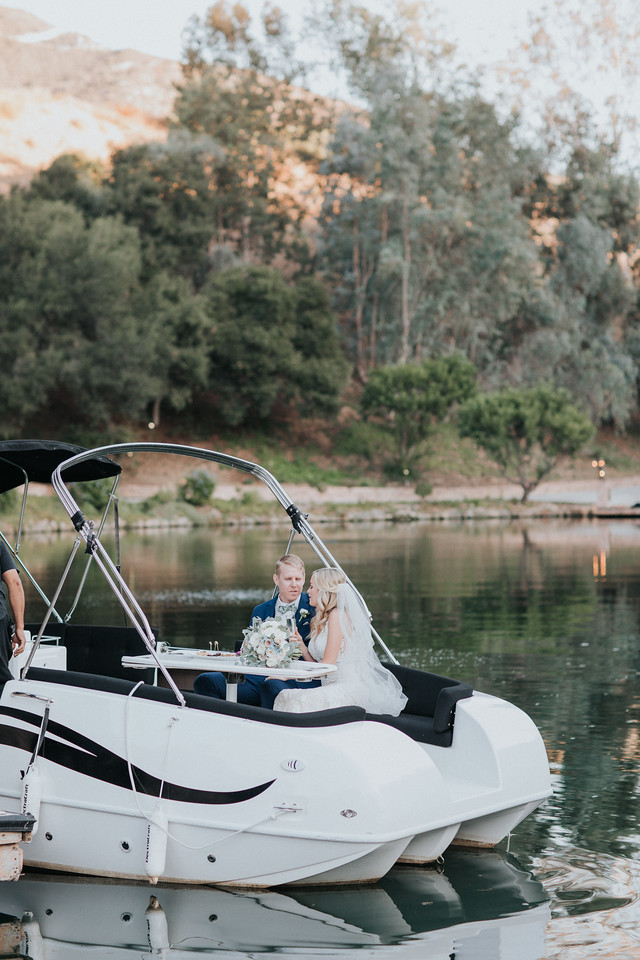 ceremony 238 X2 A bride and groom sit together on a docked white boat by a calm lake, surrounded by trees and hills in the background.