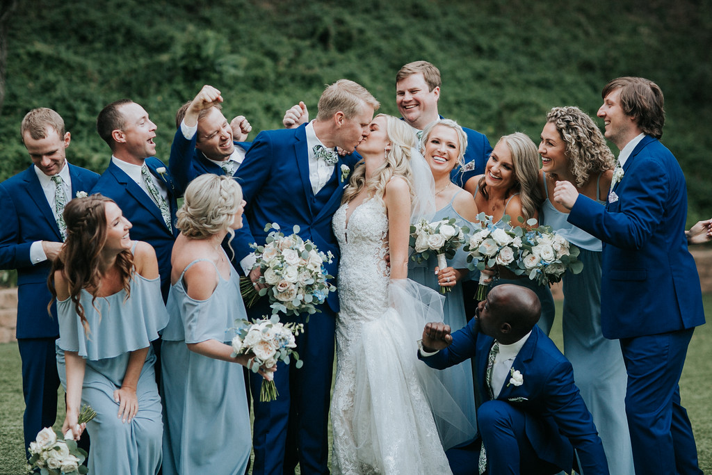 postceremony 22 X3 A bride and groom kiss while surrounded by their wedding party, all dressed in blue attire, posing outdoors with bouquets and greenery in the background.