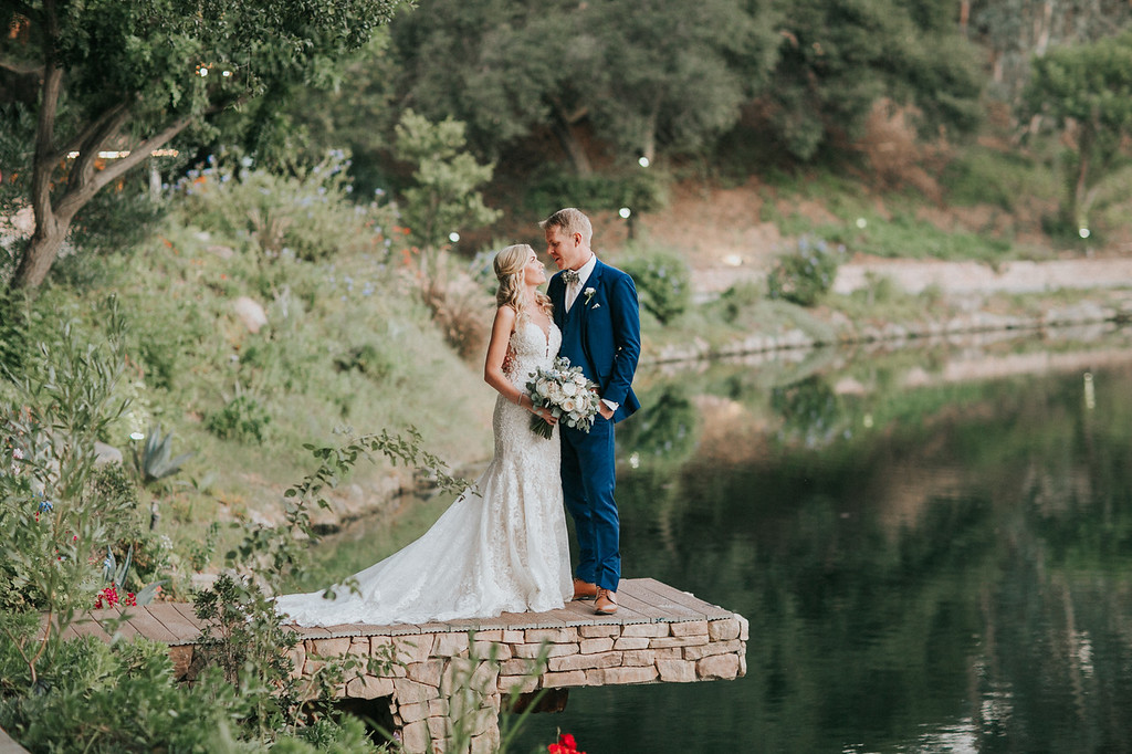 postceremony 85 X3 A bride and groom stand together on a stone dock by a lakeside, surrounded by greenery, with the bride holding a bouquet and looking at each other.