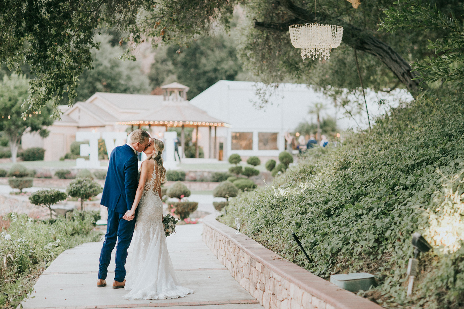 postceremony 89 X3 A bride and groom kiss on a garden pathway during an outdoor wedding, with greenery and a building in the background.