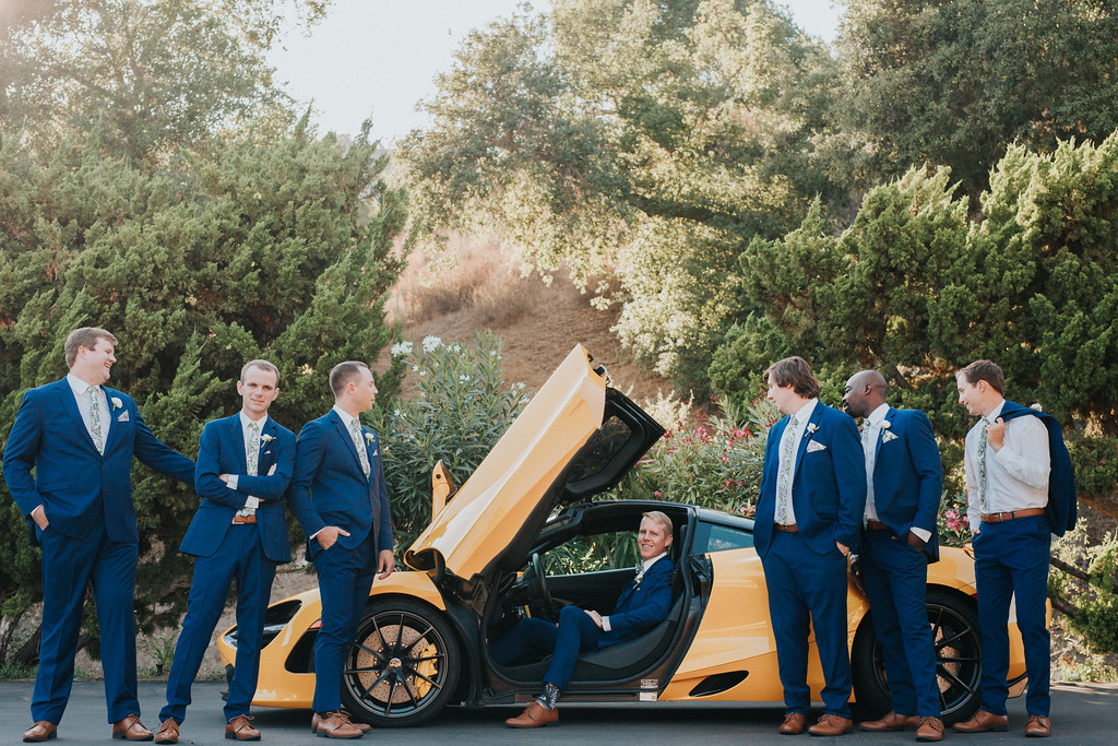 preceremony 285 X3 Six men in blue suits pose around a yellow sports car with its door open; one man sits inside the car while the others stand nearby, outdoors with trees in the background.