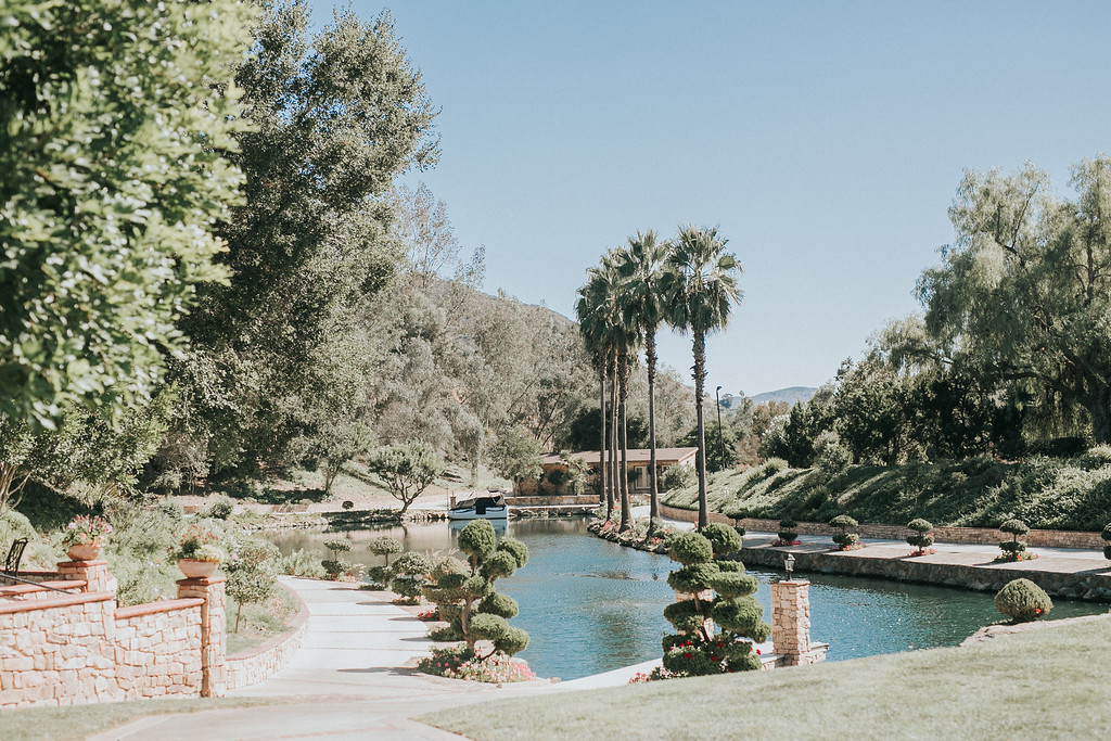 preceremony 3 X3 A landscaped garden features a small river lined with manicured trees, palm trees, stone walls, and a clear blue sky in the background.