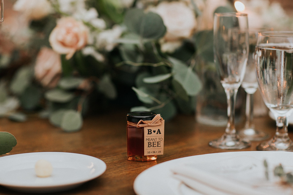 reception 17 X3 A small jar of honey with a "Meant to Bee" label sits on a decorated table with plates, glasses, and floral greenery in the background.