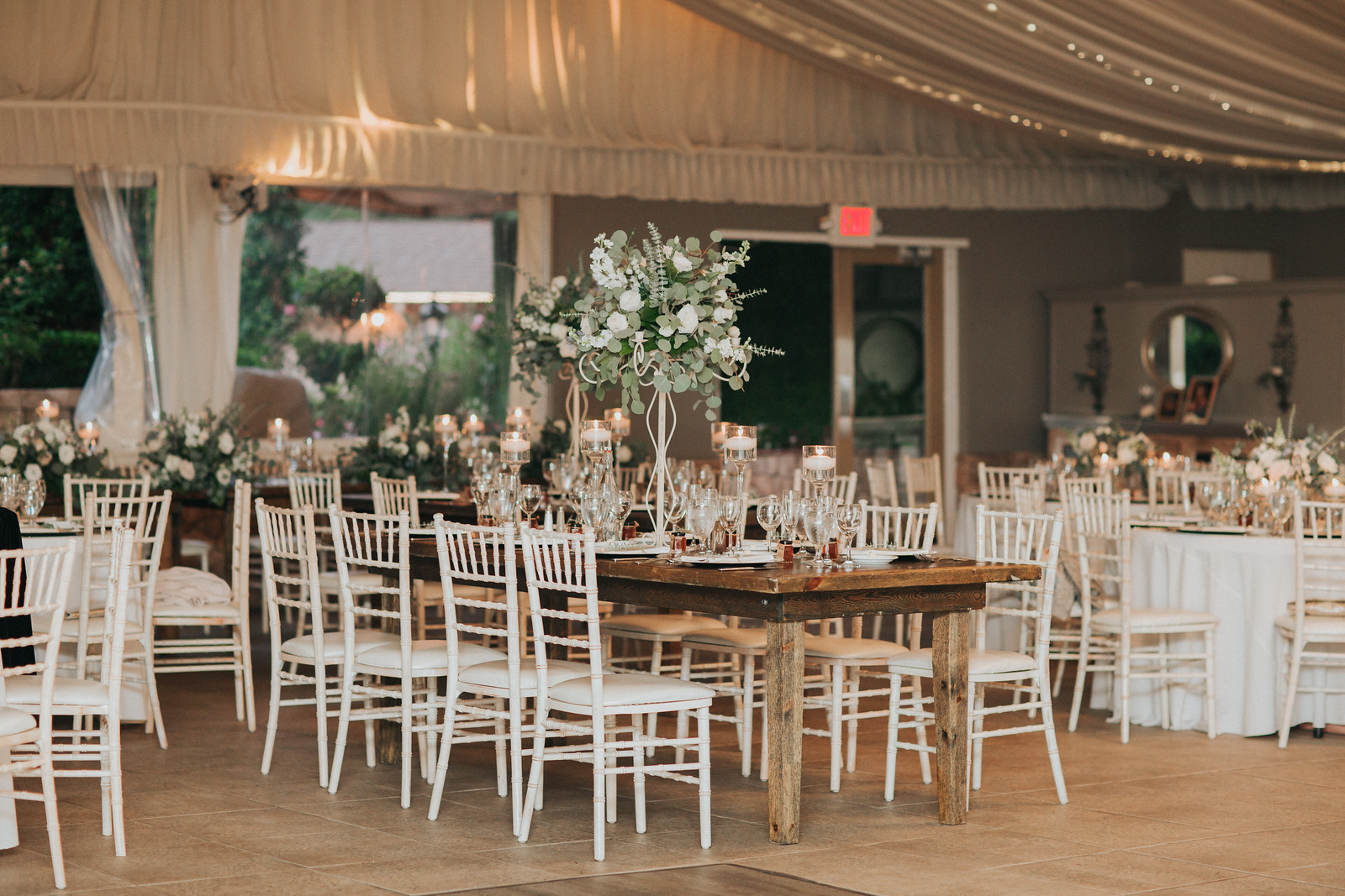 reception 2 X3 Elegant indoor wedding reception setup with white chairs, round tables, floral centerpieces, candles, and draped ceiling lights.