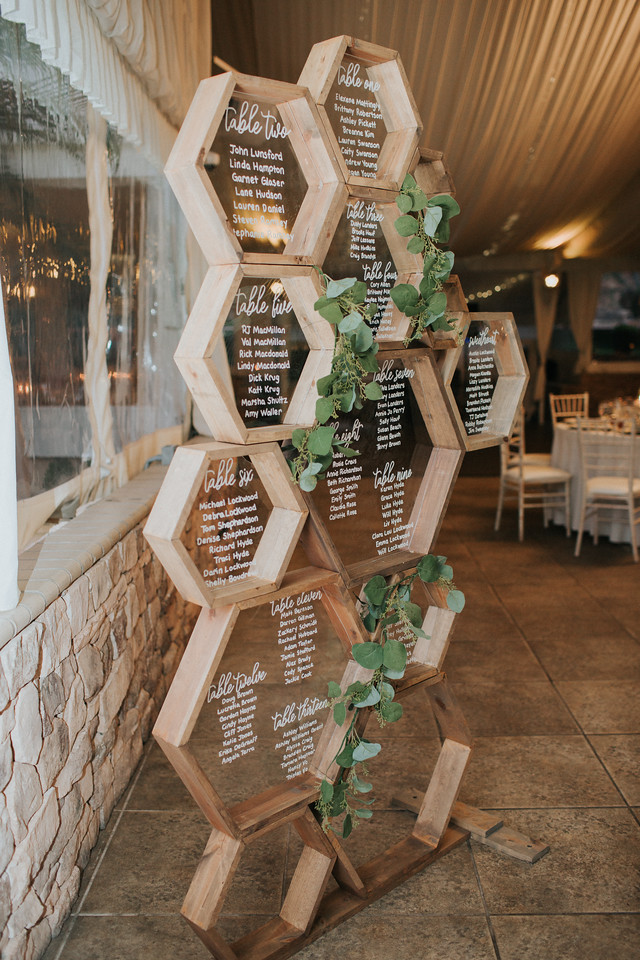 reception 4 X2 A wooden hexagonal seating chart display lists table assignments, with white lettering and green foliage accents, inside an event venue.