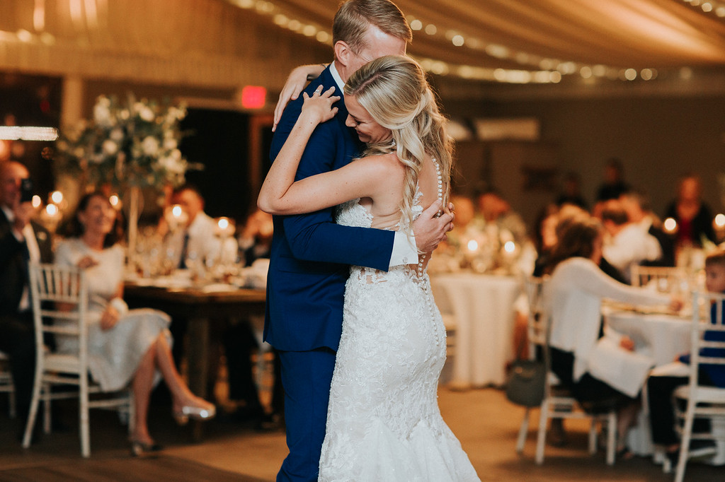 reception 41 X3 A bride and groom share a first dance in a warmly lit reception hall with guests seated at decorated tables in the background.