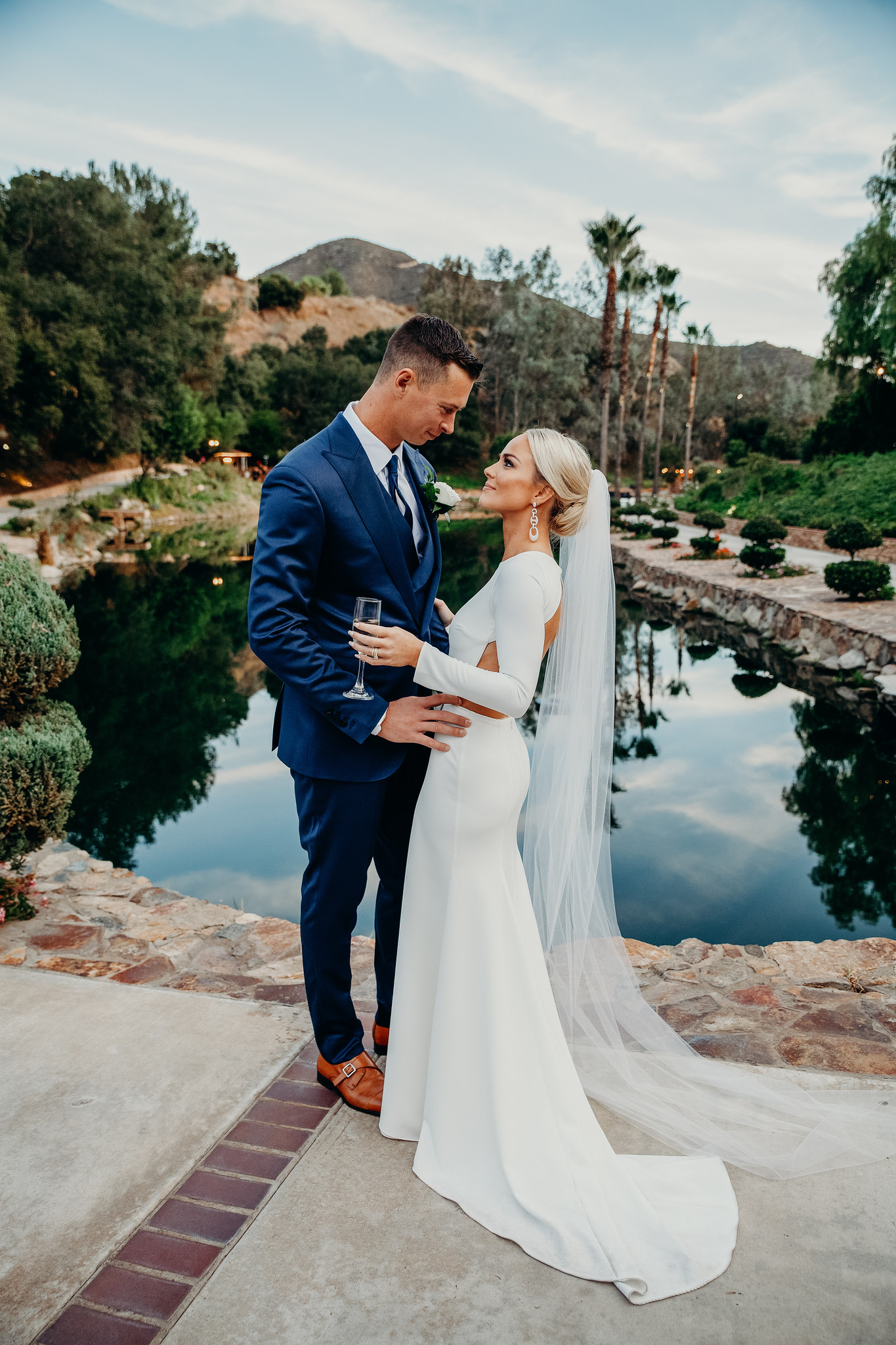 A bride and groom stand together by a pond, gazing at each other. The bride wears a white dress and veil, and the groom is in a blue suit. Lush greenery and hills are in the background.