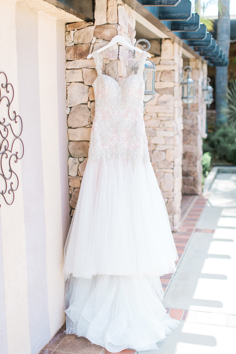 AllysonandSean1 X3 A white lace wedding gown with a tulle skirt hangs on a wooden hanger against a stone wall in an outdoor setting.