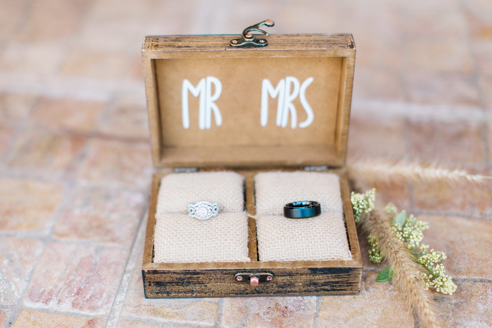 AllysonandSean21 X3 A wooden box labeled "MR" and "MRS" holds a diamond engagement ring and a black wedding band, placed on burlap cushions, on a tiled surface.