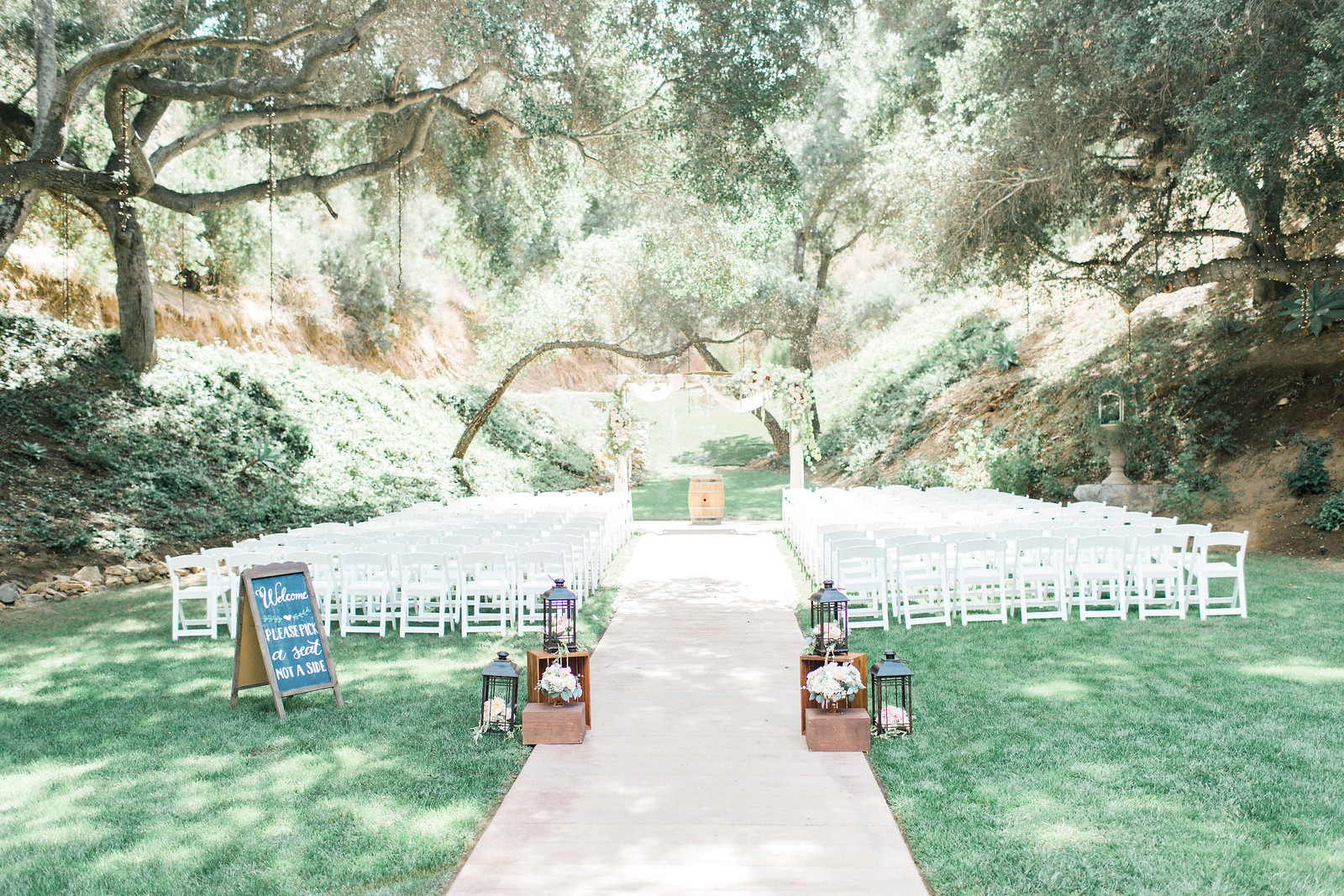AllysonandSean339 X3 Outdoor wedding ceremony setup with rows of white chairs facing an altar under large trees, decorated with lanterns and a welcome sign by the entrance.