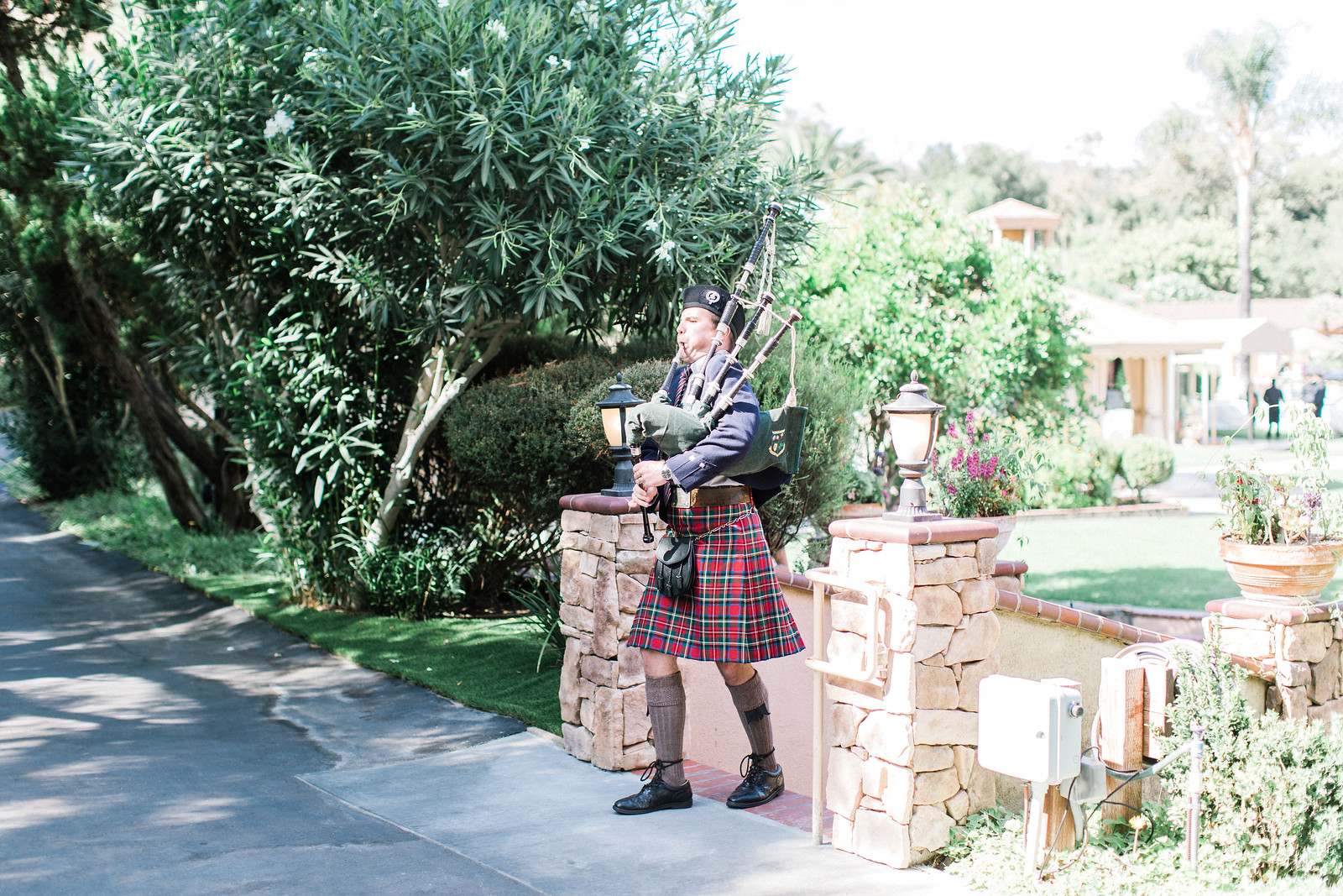 AllysonandSean355 X3 A man wearing a traditional Scottish kilt plays the bagpipes outdoors near a stone wall and garden.
