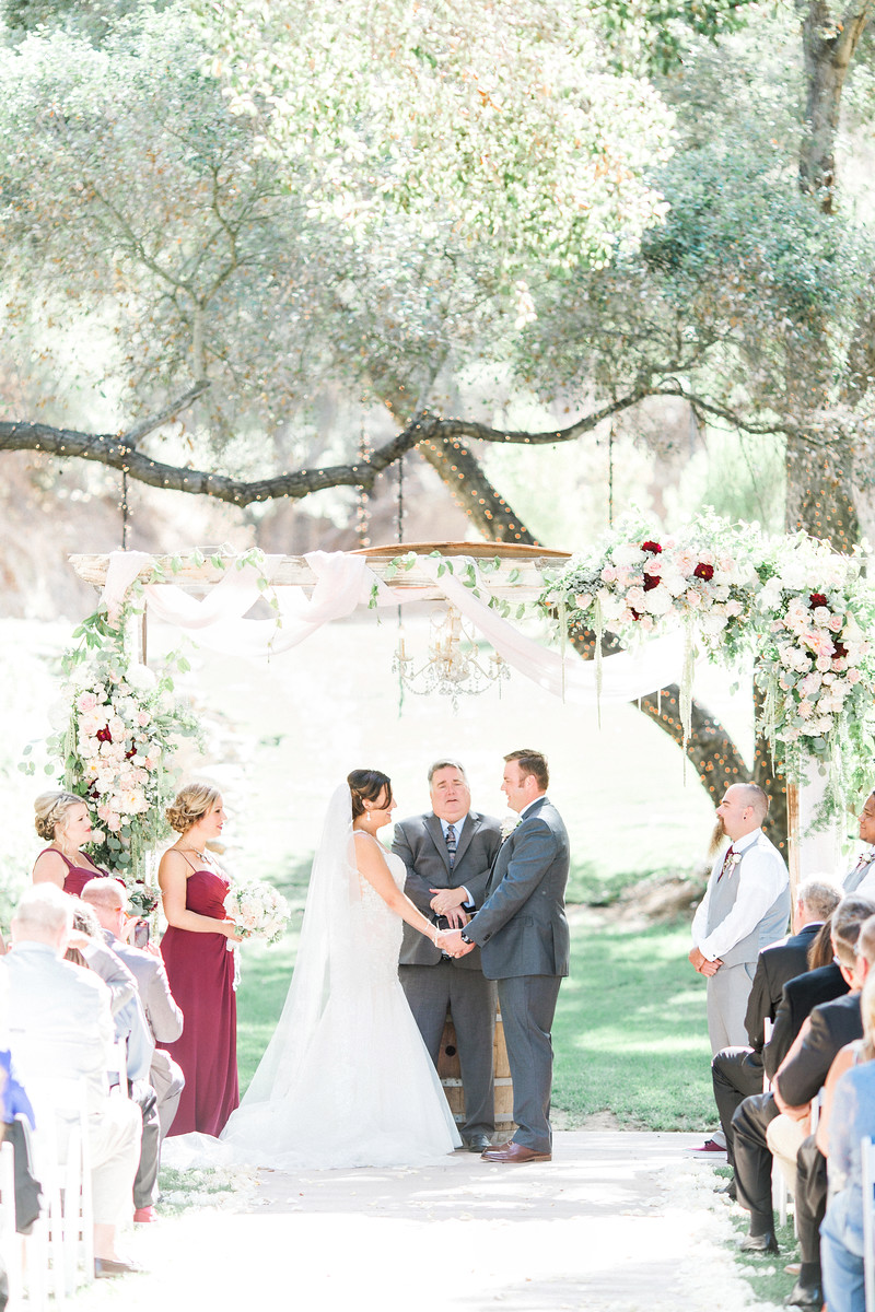 AllysonandSean386 X3 A bride and groom stand at an outdoor wedding altar, holding hands as a wedding officiant speaks; bridesmaids and groomsmen stand nearby, with guests seated in the foreground.