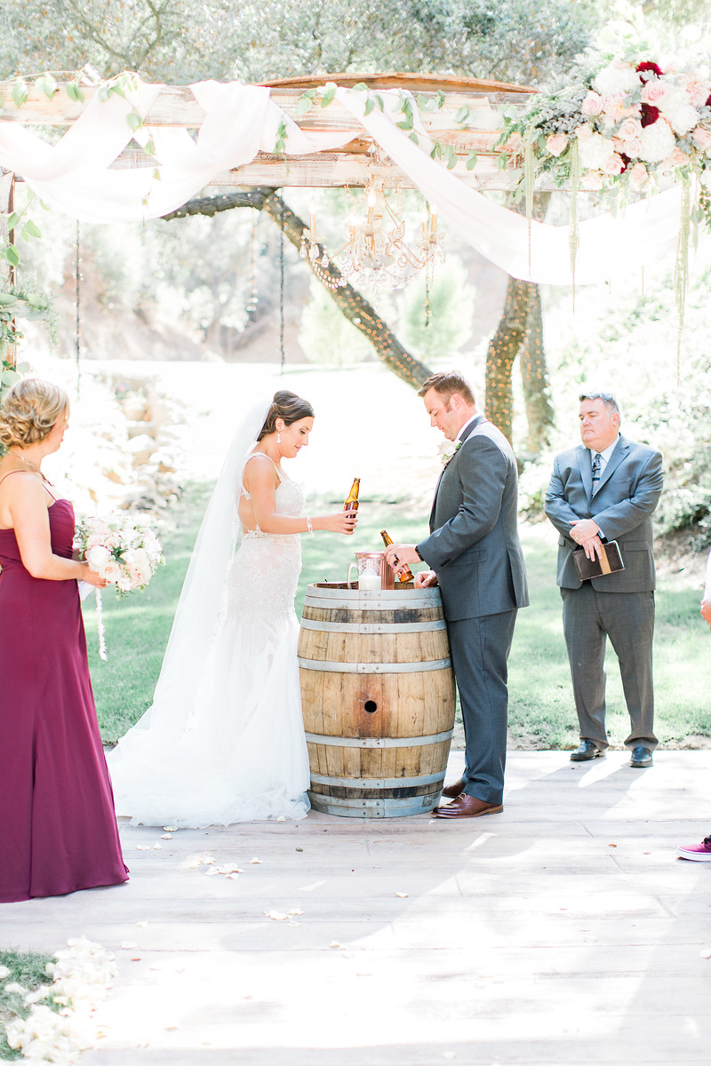 AllysonandSean451 X3 A bride and groom stand at a wine barrel during an outdoor wedding ceremony, with a bridesmaid and officiant nearby under a floral arch.