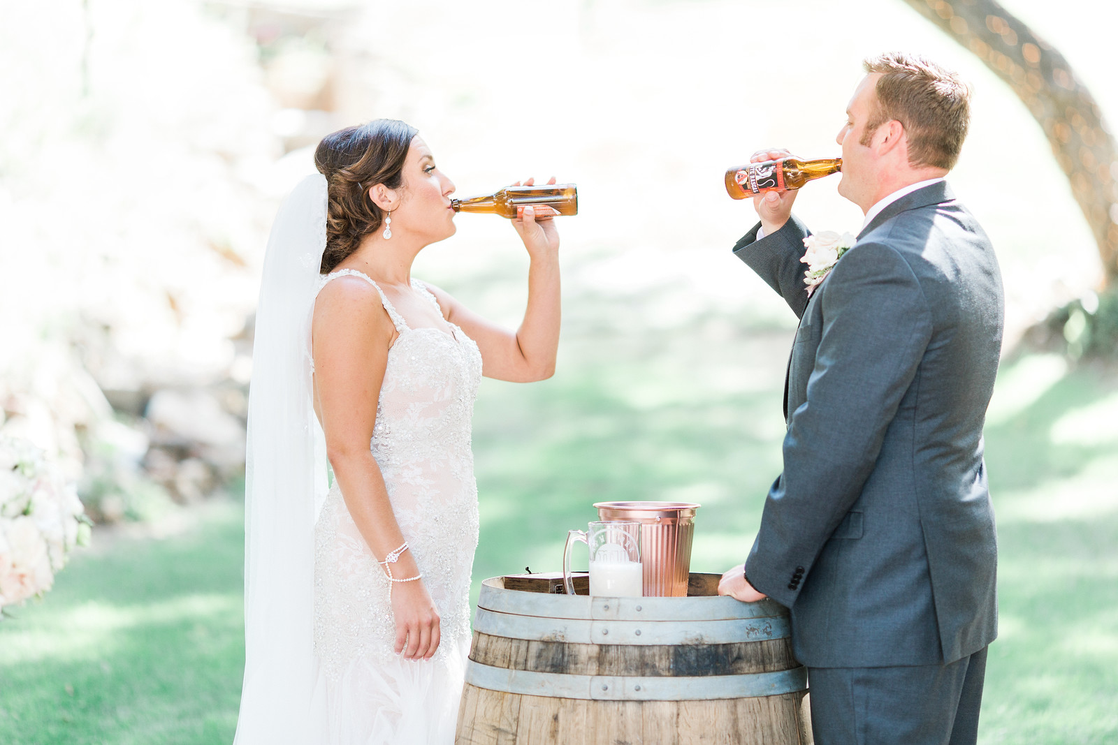 AllysonandSean452 X3 A bride and groom in wedding attire stand outdoors by a wooden barrel, each drinking from a beer bottle.