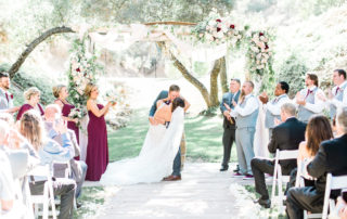 A bride and groom kiss under a flower-decorated arch at an outdoor wedding ceremony, surrounded by their bridal party and seated guests.