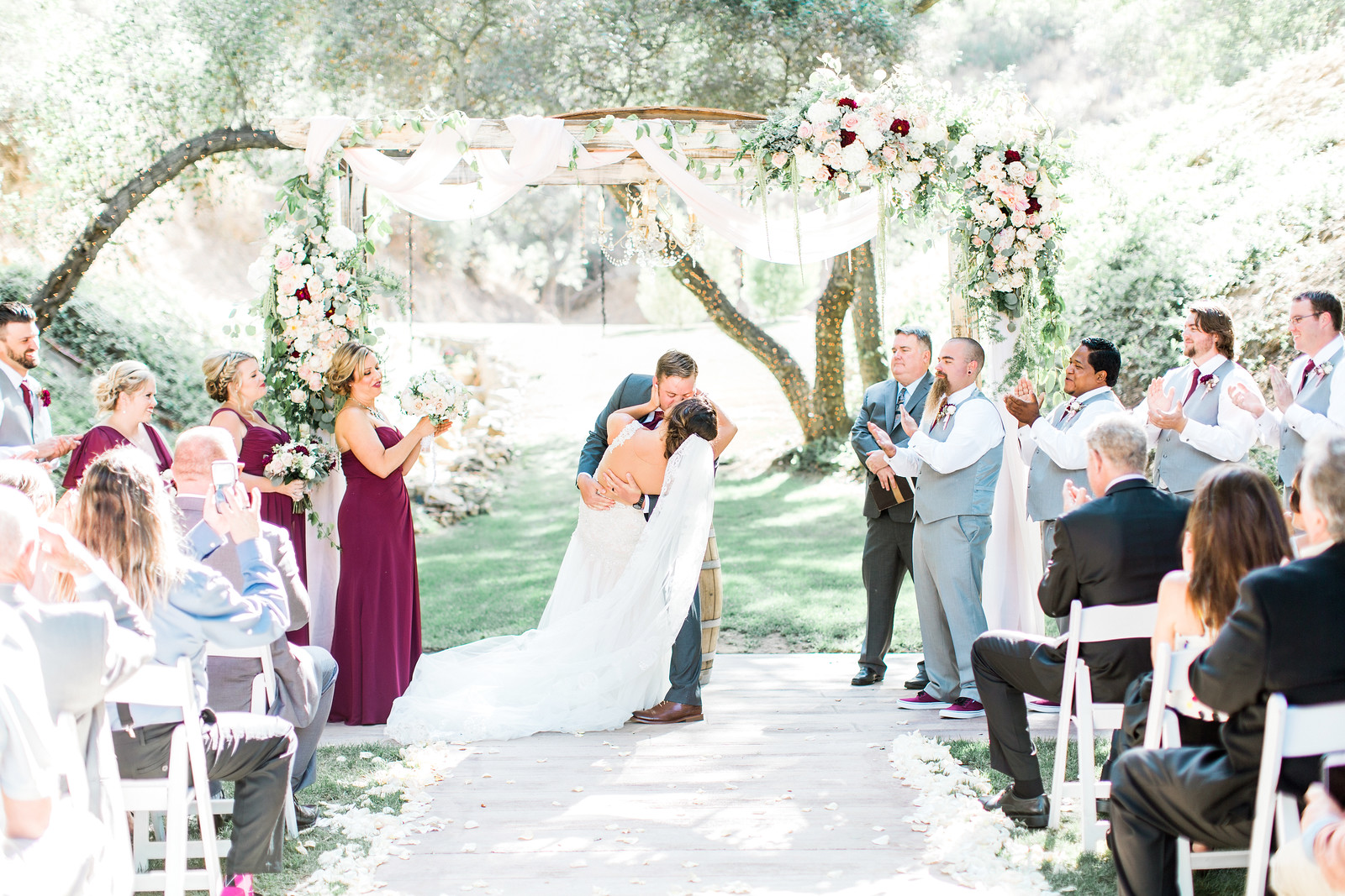 A bride and groom kiss under a flower-decorated arch at an outdoor wedding ceremony, surrounded by their bridal party and seated guests.