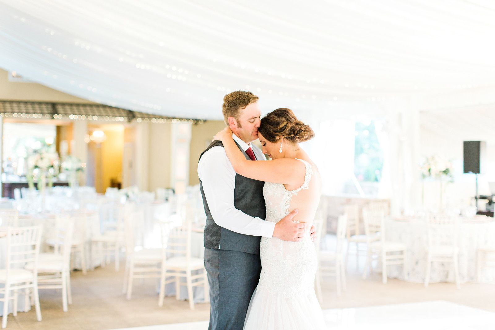 AllysonandSean608 X3 A bride and groom embrace on the dance floor in a bright, elegant wedding reception venue with white decor and empty chairs.