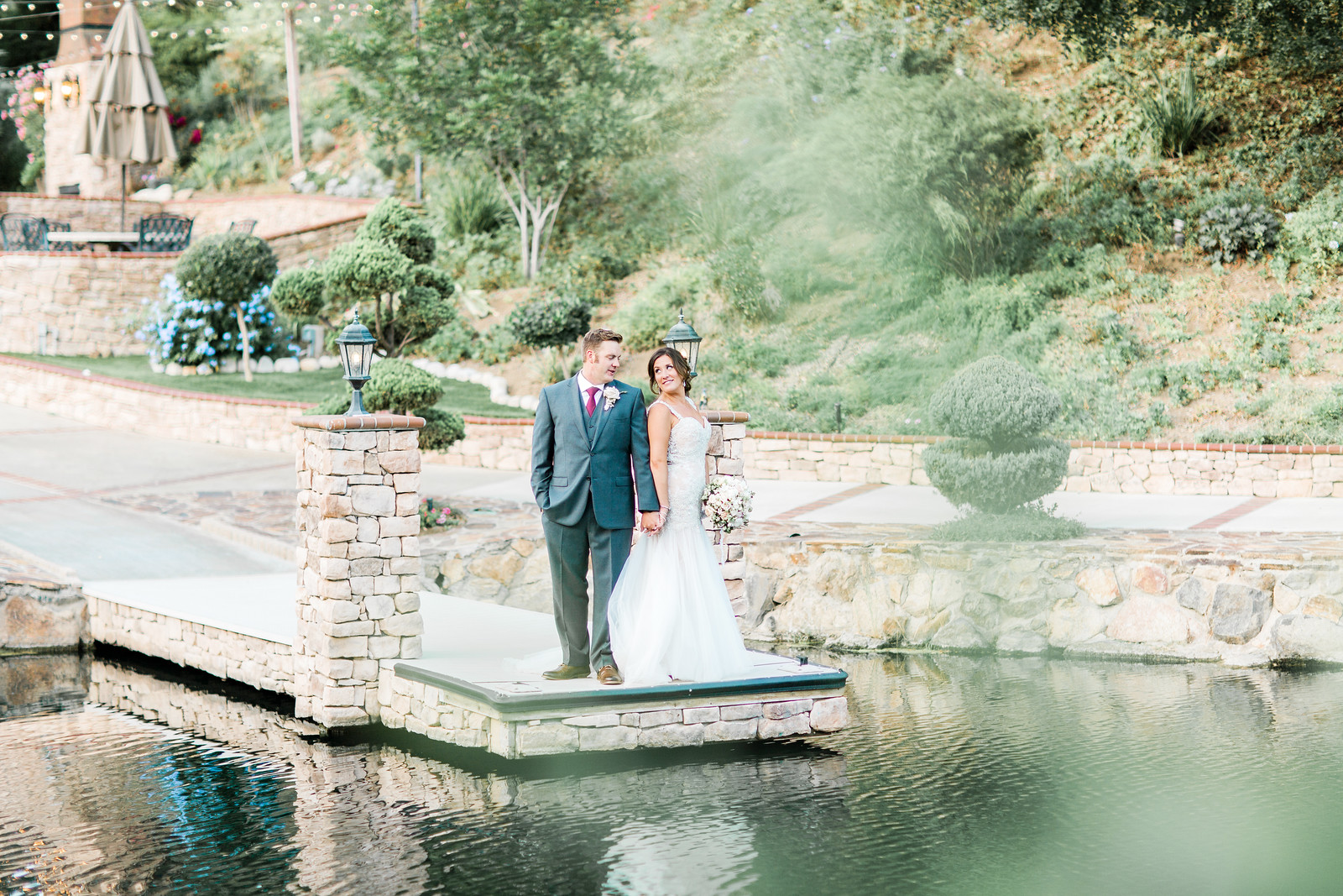AllysonandSean632 X3 A bride and groom stand together on a stone dock over a pond, surrounded by greenery and landscaped gardens.