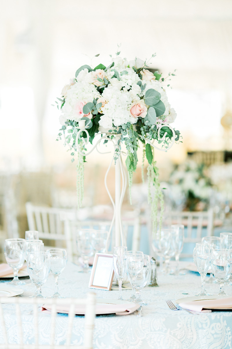 AllysonandSean665 X3 Tall floral centerpiece with white and pink flowers and greenery on a round table set with glassware, plates, and silver chairs in a bright, elegant event space.
