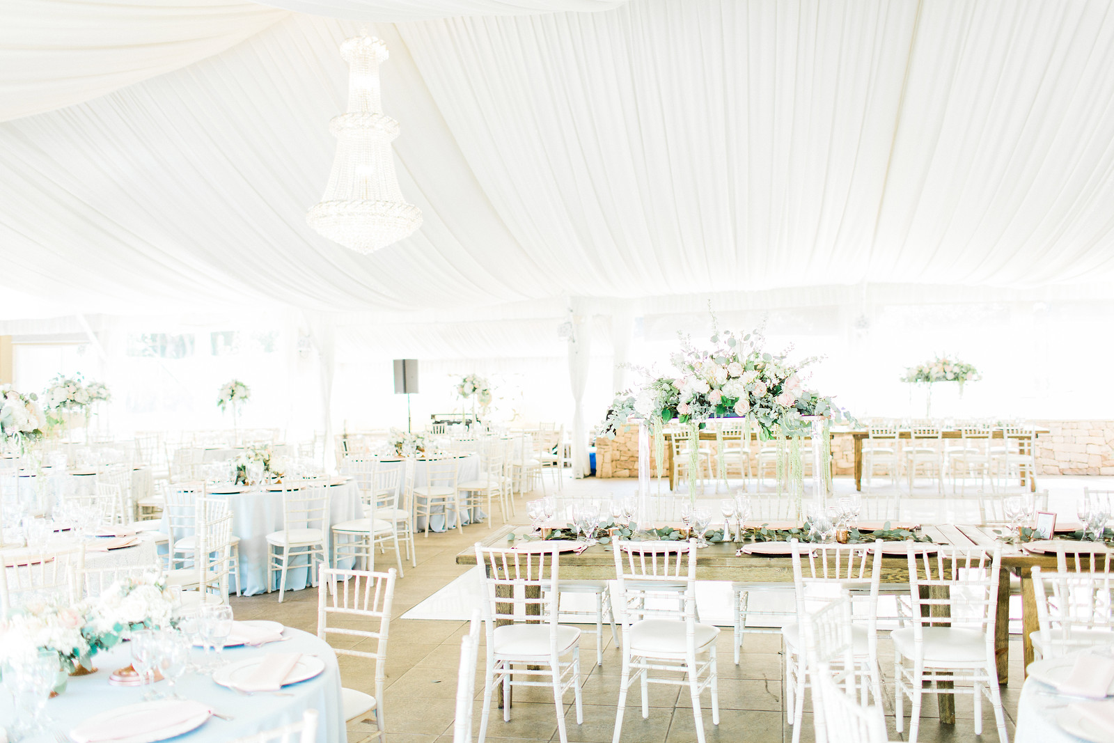 AllysonandSean666 X3 A bright, elegant wedding reception venue with white draped ceiling, chandeliers, white chairs, round tables, and floral centerpieces.