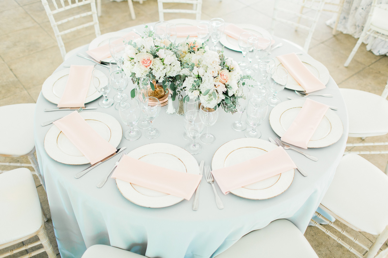 AllysonandSean667 X3 A round table set for eight with white plates, pink napkins, glassware, silver cutlery, and floral centerpieces on a light blue tablecloth.