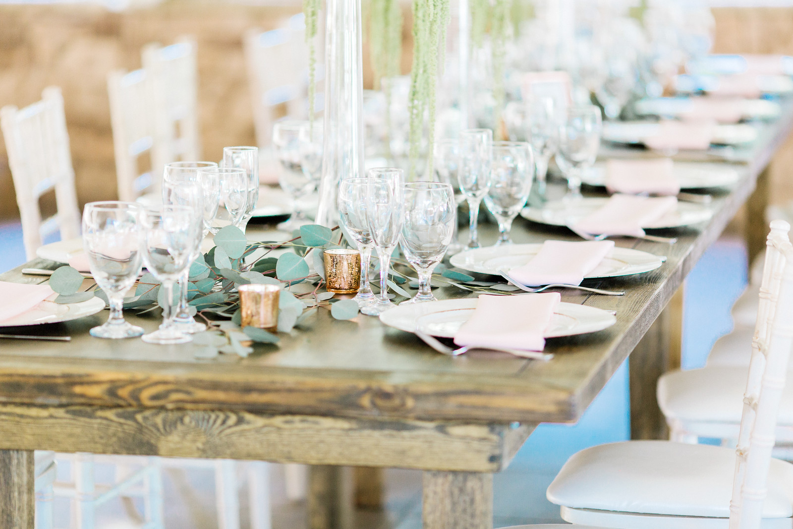 AllysonandSean681 X3 A wooden dining table is set with glassware, white plates, pink napkins, greenery, and candles, with white chairs arranged along both sides.
