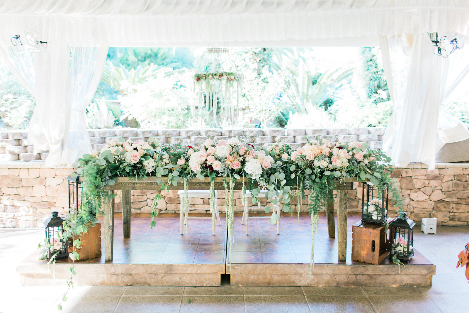 AllysonandSean704 X3 A wooden table decorated with pink and white flowers and greenery, flanked by lanterns, set under a white canopy with stone walls and a garden in the background.
