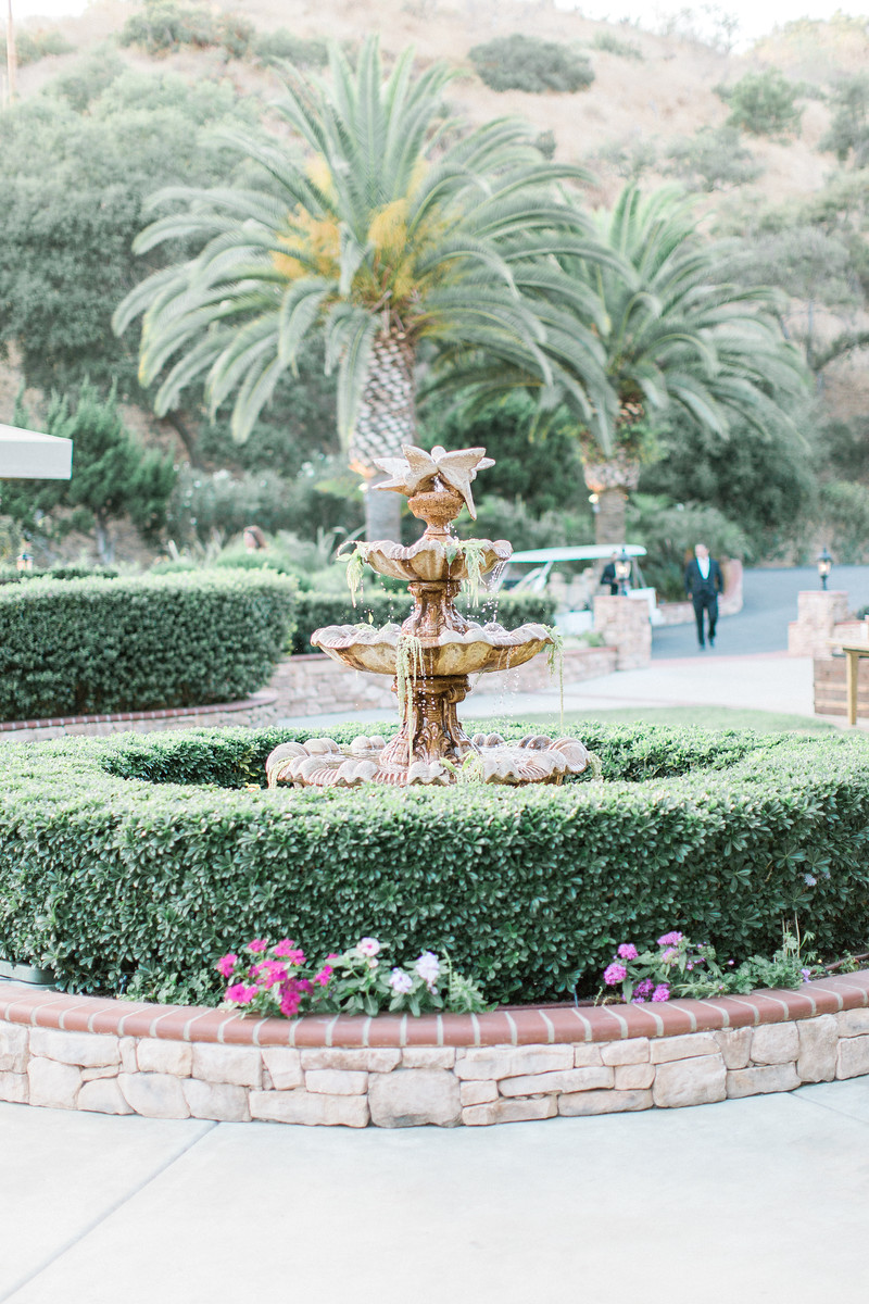 AllysonandSean731 X3 A tiered stone fountain surrounded by trimmed hedges and flowers, with palm trees and a person walking in the background.