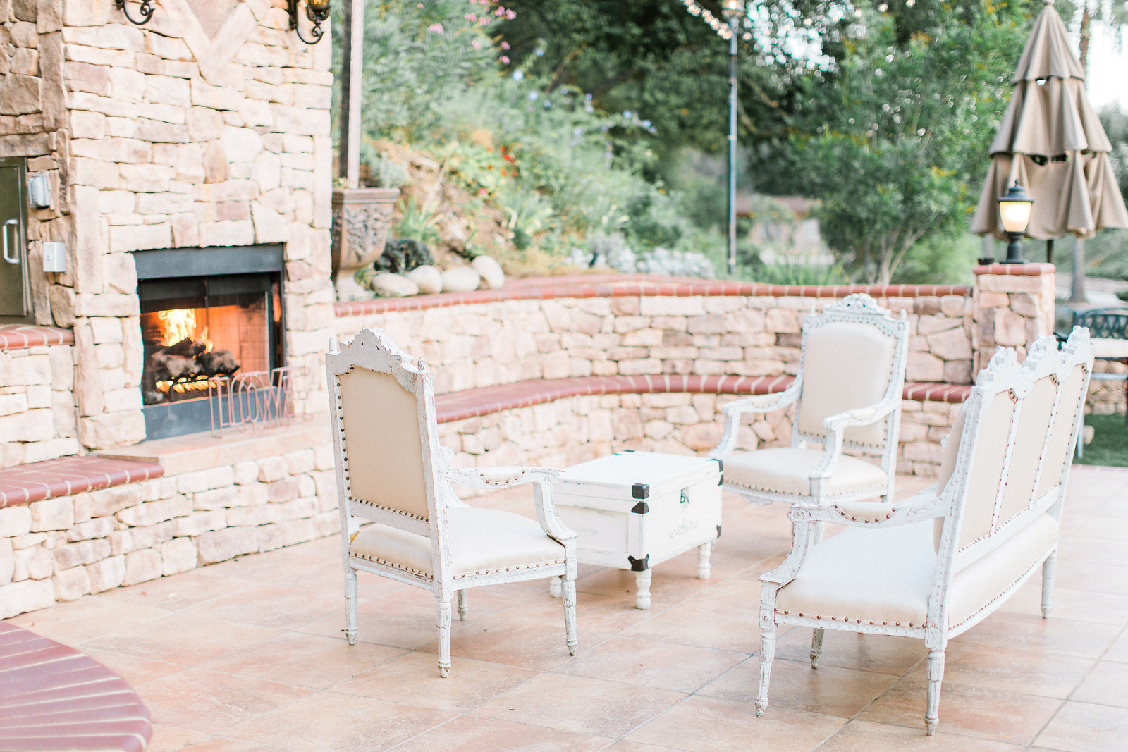 AllysonandSean734 X3 Outdoor patio with white vintage-style chairs and a matching table arranged around a lit stone fireplace, surrounded by stone walls and greenery.