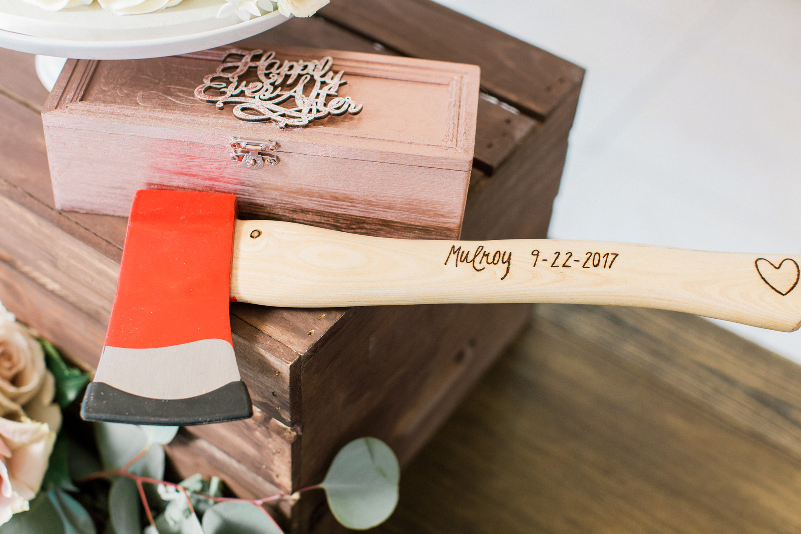 AllysonandSean758 X3 A wooden-handled axe with "Mulroy 9-22-2017" engraved on it rests on a wooden box, with flowers nearby and a "Happily Ever After" sign in the background.