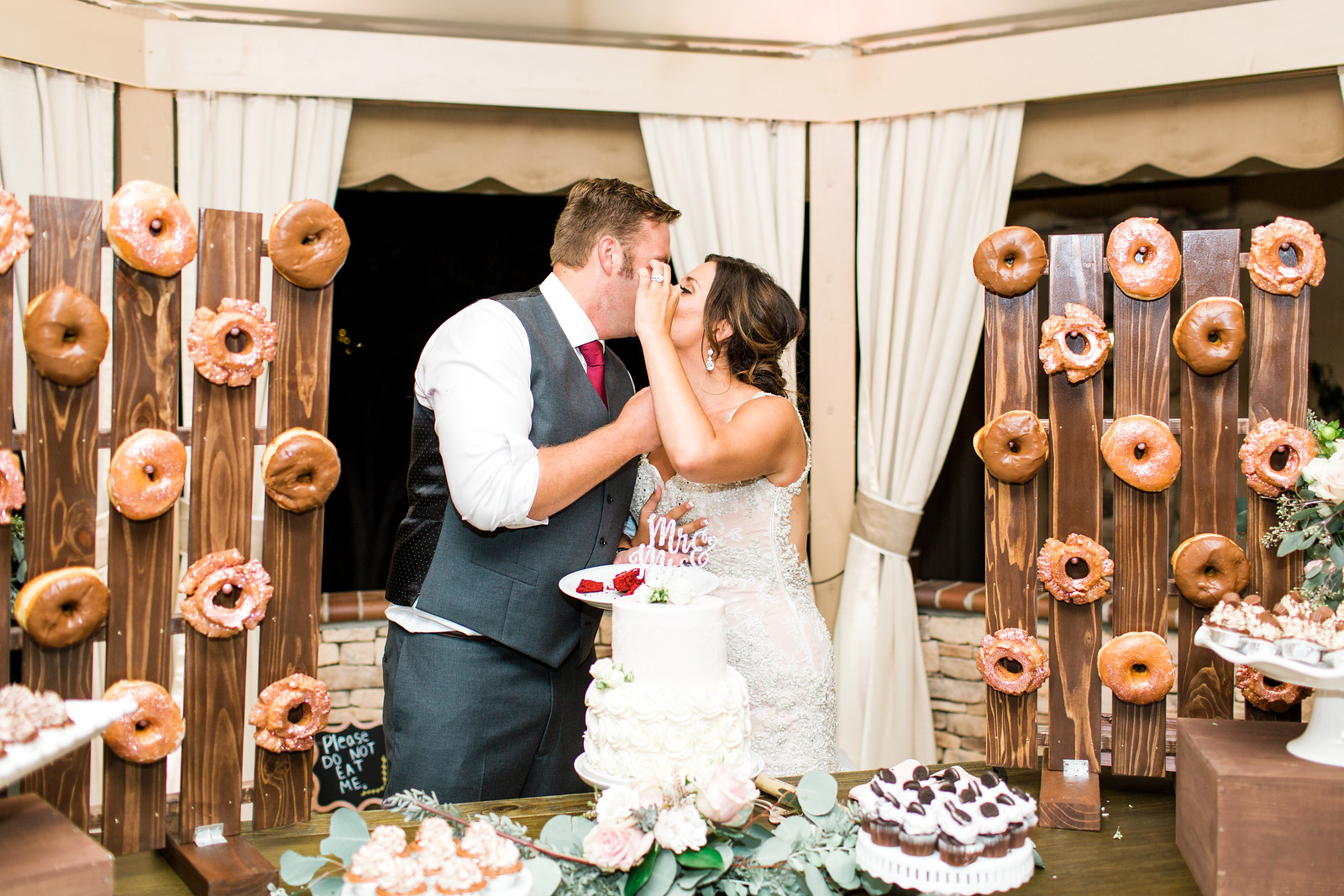 AllysonandSean901 X3 A bride and groom kiss in front of a wedding cake, surrounded by wooden stands displaying assorted donuts and desserts.