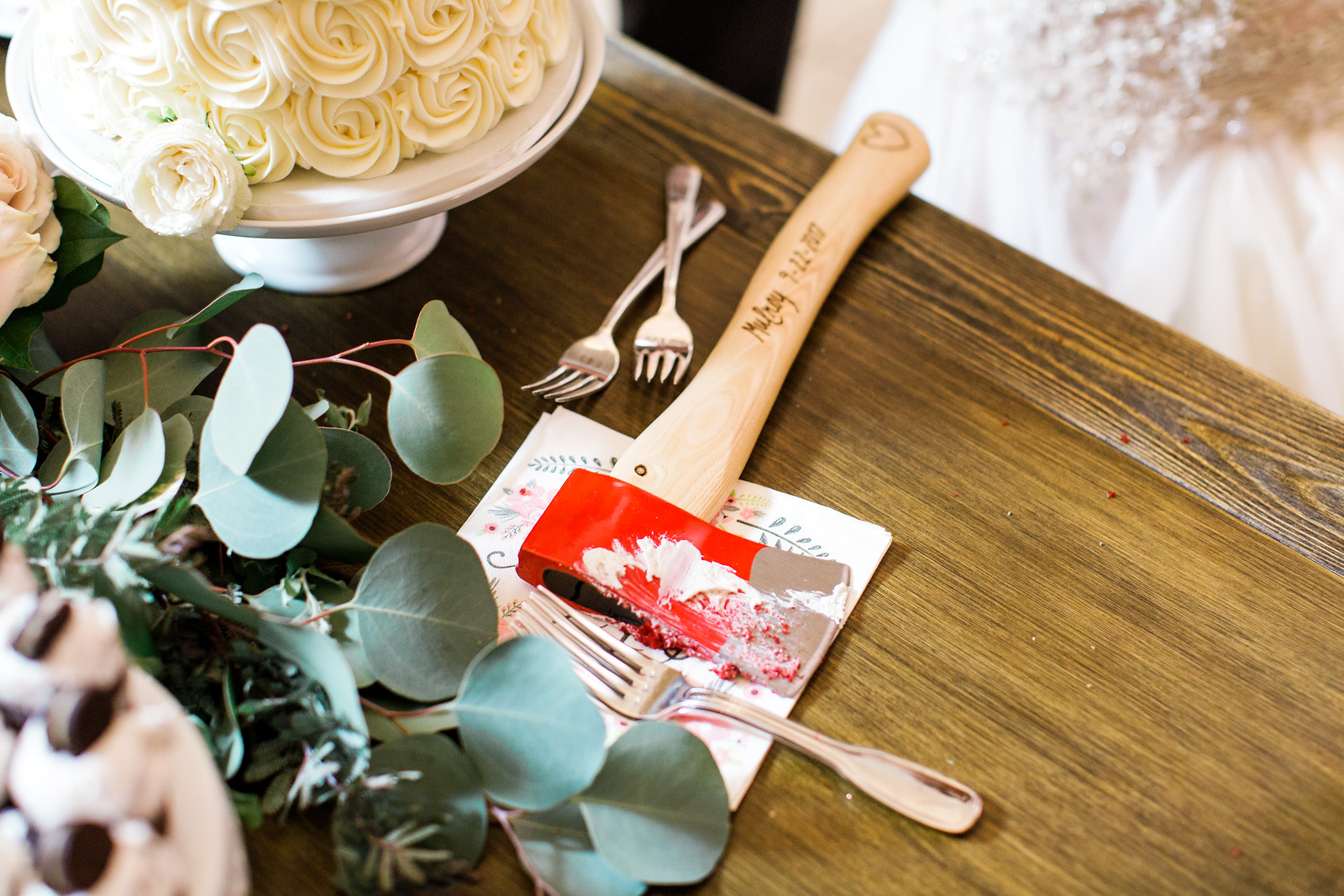 AllysonandSean908 X3 A cake-cutting hatchet with red and white icing sits on a napkin next to a wedding cake and greenery on a wooden table.