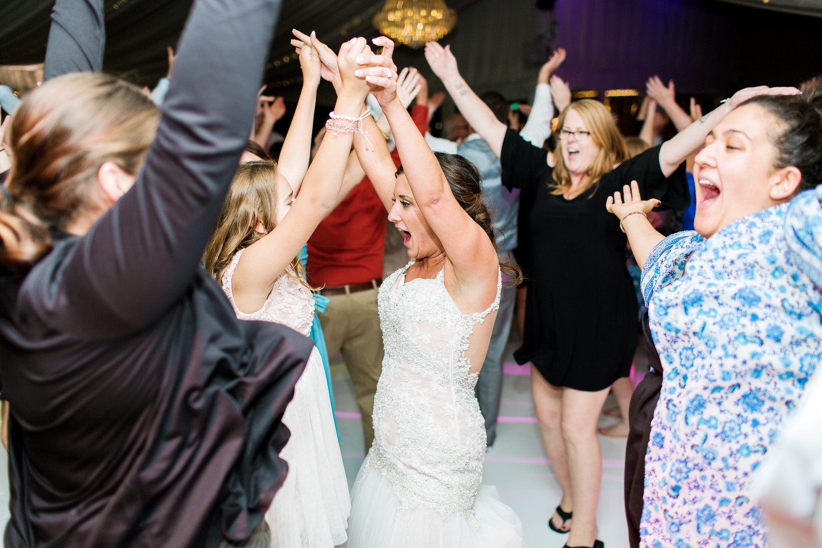 AllysonandSean964 X3 A bride in a white dress dances with guests at a wedding reception, surrounded by people with raised arms and joyful expressions.