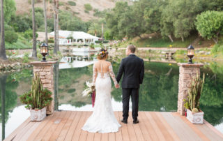 A bride and groom stand hand in hand on a wooden dock by a calm lake, facing away, surrounded by greenery and stone pillars with potted plants.