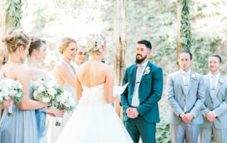 A bride reads vows to the groom during an outdoor wedding ceremony; bridesmaids and groomsmen stand beside the couple holding bouquets.
