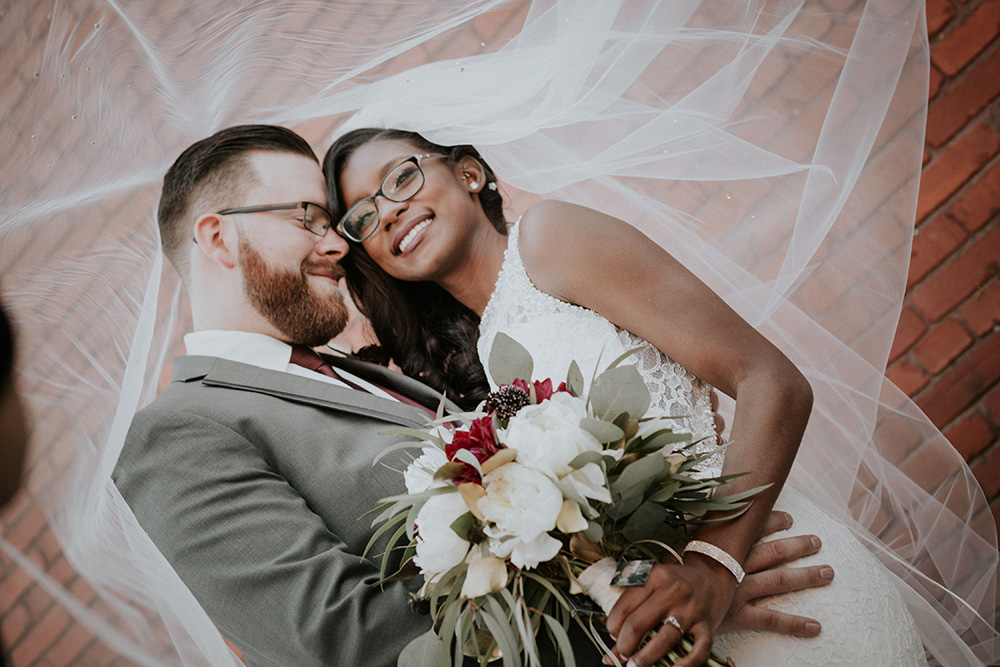 A bride and groom embrace, smiling under a flowing veil, with the bride holding a bouquet of white and burgundy flowers against a brick wall background.