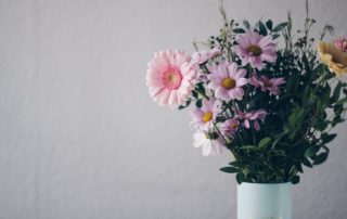 A bouquet of pink and yellow daisy-like flowers with green leaves arranged in a light blue vase against a plain light background.