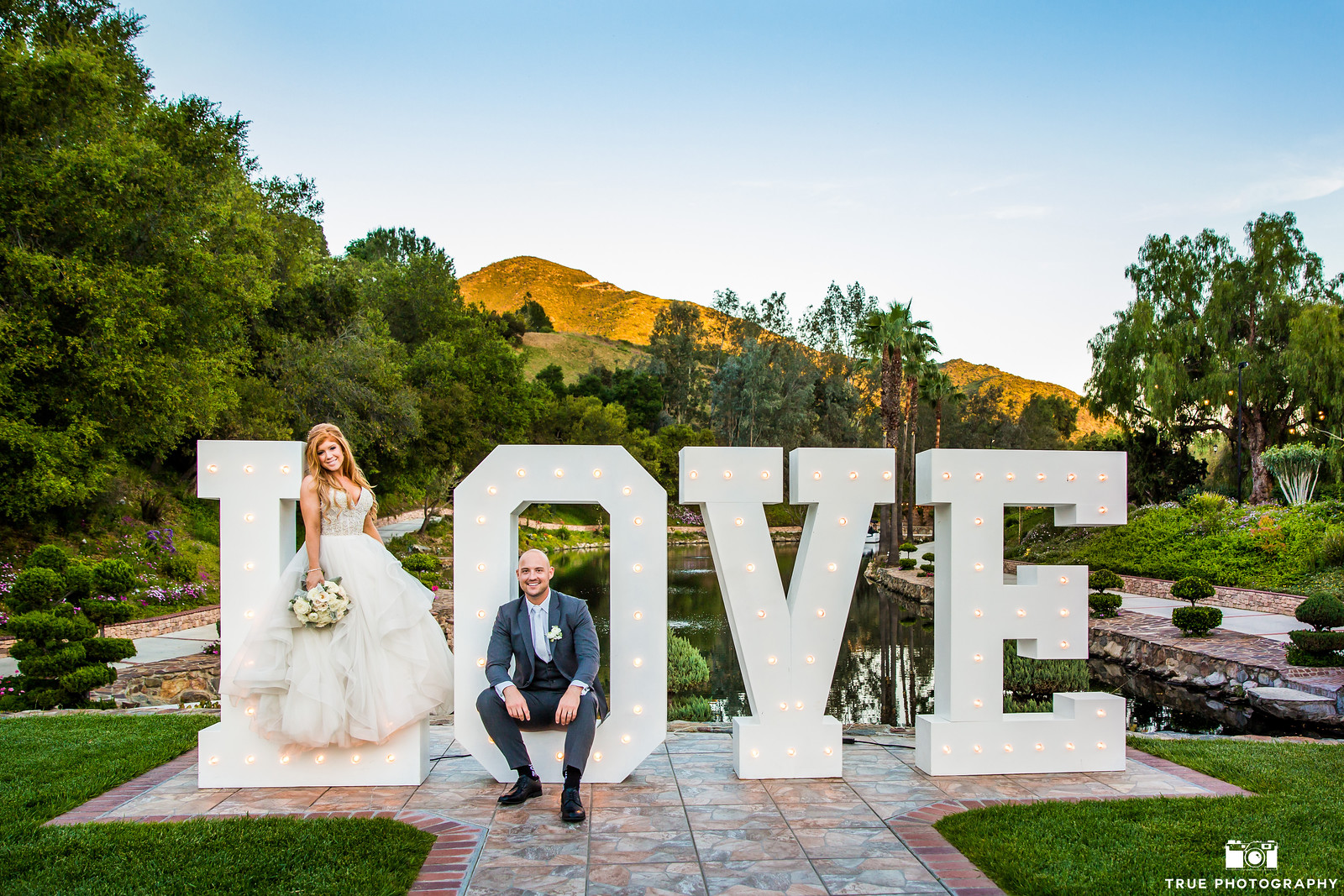 0001Kaitlyn Brian X3 A bride stands on the "L" and a groom sits on the "V" of large, lit-up "LOVE" letters outdoors, with greenery and mountains in the background.