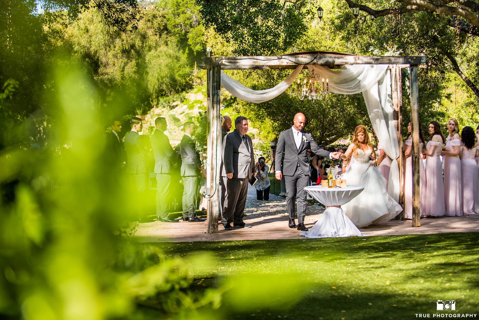 0058Kaitlyn Brian 1 X3 A bride and groom stand under a decorated canopy during an outdoor wedding ceremony, with guests and bridesmaids in the background.