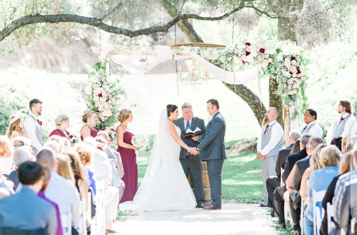 A bride and groom stand facing each other at an outdoor wedding ceremony under a floral arch, surrounded by their wedding party and guests.