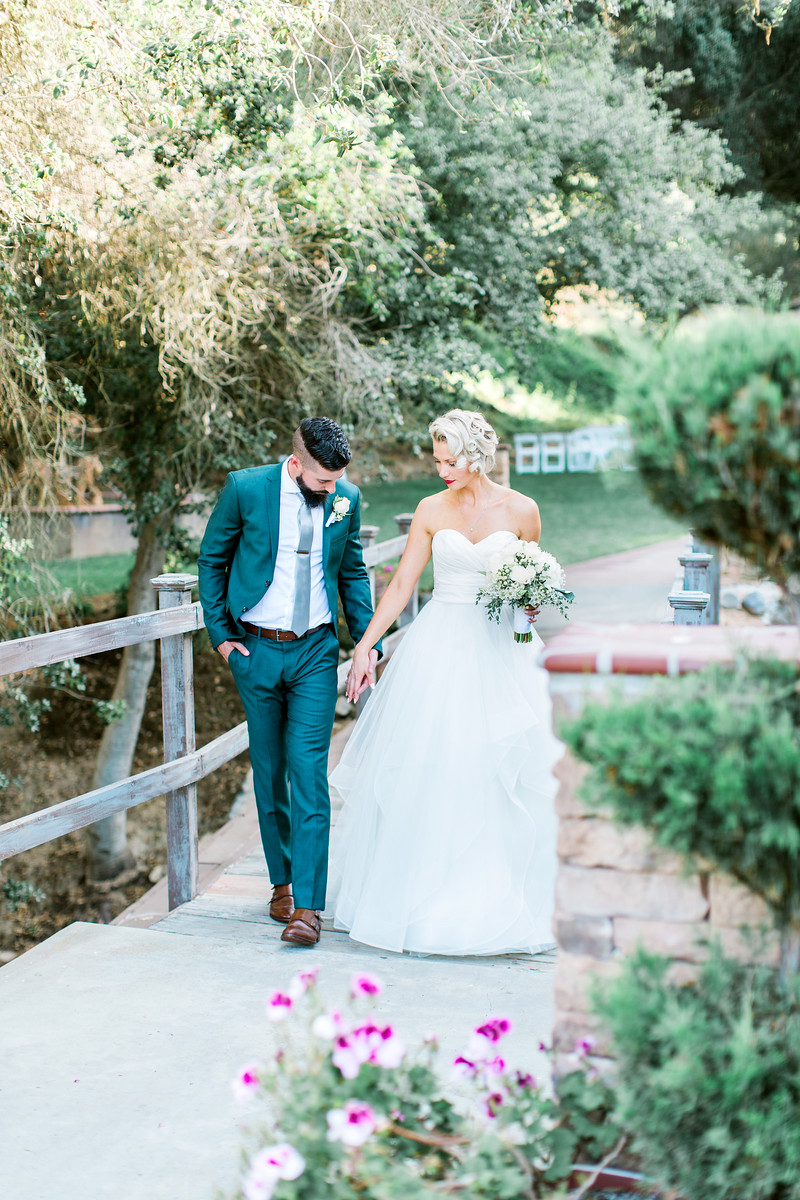 A bride in a white gown and a groom in a blue suit walk hand in hand on a garden bridge surrounded by greenery and flowers.