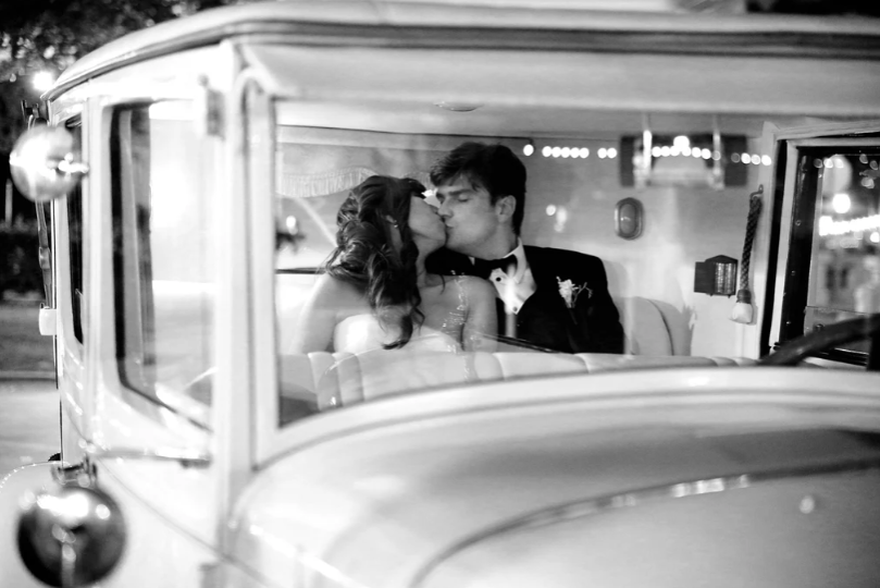 A couple dressed formally kisses while sitting in the front seat of a vintage car at night.