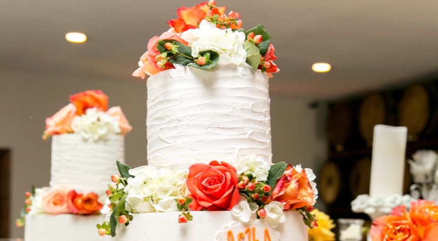 Two-tiered white cake decorated with textured frosting and adorned with red, white, and orange flowers, displayed indoors under warm lighting.