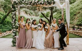 A bride and bridesmaids in pastel dresses and one groomsman pose playfully under a floral wedding arch in an outdoor garden setting.