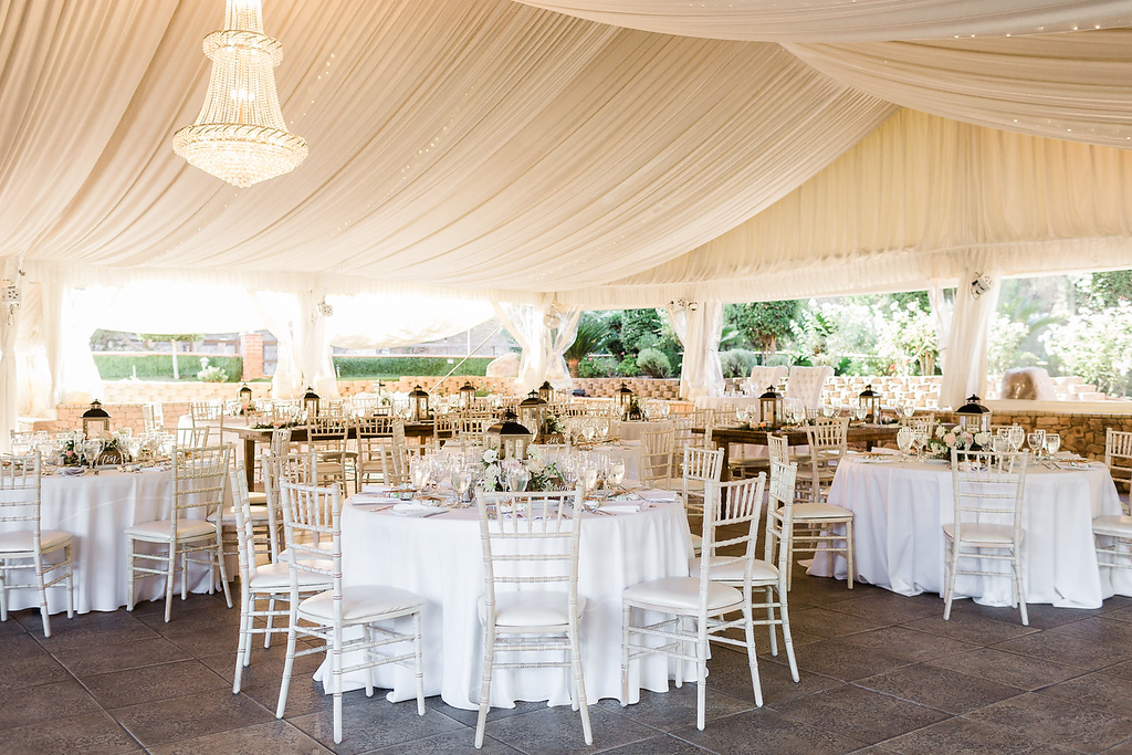 A decorated event venue with round tables covered in white tablecloths, white chairs, glassware, and a chandelier hanging from a draped ceiling.